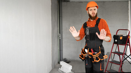 Male bearded builder in hard hat and blue overalls with working tools on belt showing two hand gesture no thank or stop. repair, construction and building
