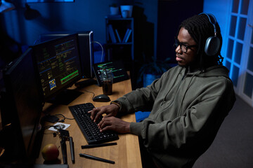 Young Black man wearing headphones sitting at desk typing on computer keyboard, multiple monitors displaying code, focused on programming in workspace at night