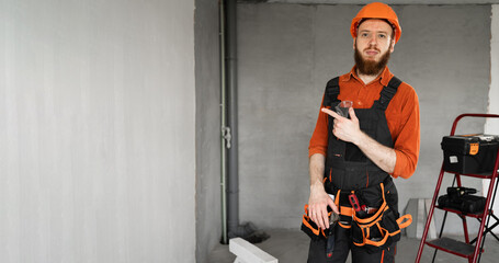 Male bearded builder in hard hat and blue overalls with working tools on belt point index finger aside on area. Repair, construction and building