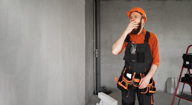 Tired lazy Male bearded builder in hard hat and blue overalls with working tools on belt sleepy yawning after overworked. Repair, construction and building