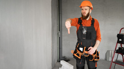 Unhappy Male bearded builder in hard hat and blue overalls with working tools on belt showing...
