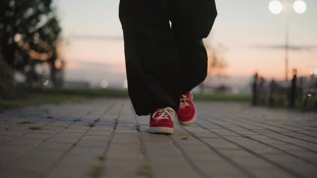 dancer feet on pavement at dusk, red sneakers executing shuffle steps along urban park promenade, slow motion closeup captures textured concrete, warm sunset bokeh and soft lamplight, street performer