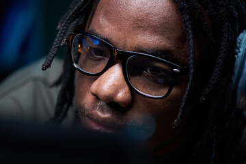 Portrait of young Black man wearing glasses focusing intently on computer screen, close up showing facial features and concentration, technology reflected in lenses