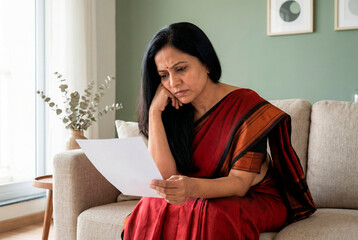 A woman with long black hair wearing a traditional Indian red sari sits on a beige couch looking concerned while reading a document.