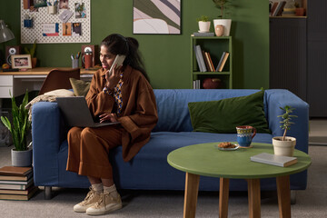 Young South Asian woman sitting on sofa multitasking with smartphone and laptop, working remotely in living room, focused on conversation while managing tasks