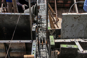 Workers pour wet concrete into a reinforced foundation formwork, showing steel rebar, wooden supports, and close-up construction details from an overhead perspective.