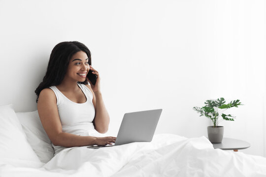 Happy african american woman freelancer talking on mobile phone and using laptop, working from home during isolation, having conversation on phone, sitting in bed and smiling, free space