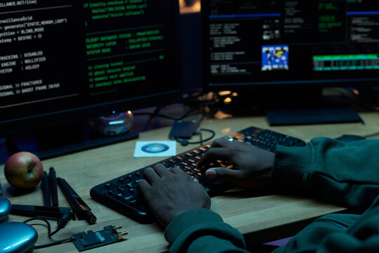 Black man typing on keyboard using desktop computer with multiple monitors displaying programming code and cybersecurity interface, hands on desk with electronic components - Powered by Adobe