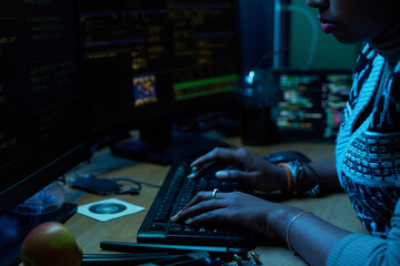 Young South Asian woman typing on computer keyboard in dark office, focusing on multiple monitors displaying code and data, hands with bracelets, working on programming task