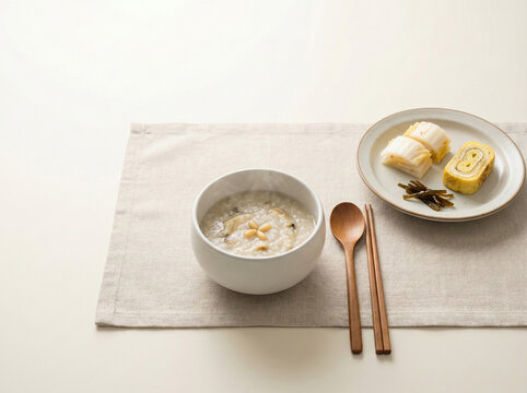Korean Abalone Porridge (Jeonbokjuk) served with pine nuts, kimchi, and rolled egg on a clean minimalist linen setting.