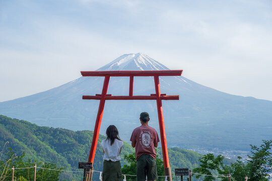 View of two people gazing at the snow-capped peak of Mount Fuji framed by a vibrant red torii gate amidst lush greenery, Fujikawaguchiko, Yamanashi, Japan.