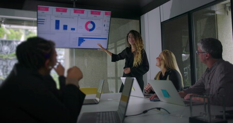 Young woman presenting data chart to colleagues while blonde businesswoman and team members listen attentively in collaborative office meeting