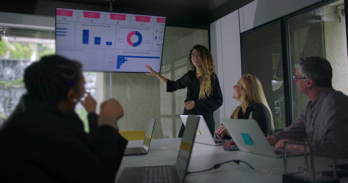 Young woman presenting data chart to colleagues while blonde businesswoman and team members listen attentively in collaborative office meeting - Powered by Adobe
