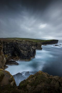 View of rugged cliffs meet the turbulent, foamy sea under a foreboding sky, a silent drama unfolding in the Scottish landscape, Kirkwall, Scotland, United Kingdom.