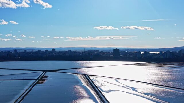 A large body of water reflects sunlight as a city skyline is seen in the distance. Clouds float in a blue sky creating a bright atmosphere during daytime.