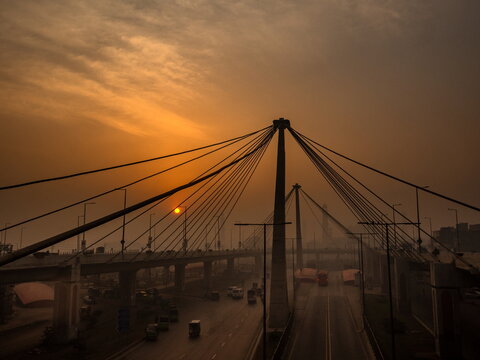 View of an orange sunset illuminating a modern cable-stayed bridge silhouetted against the sky, spanning a busy road with vehicles, Lahore, Punjab, Pakistan.