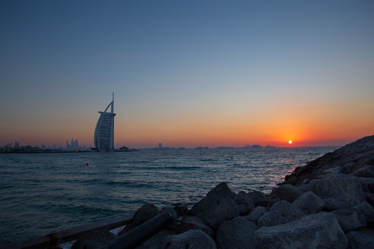 View of the radiant sun dips below the horizon, casting a warm glow over the Burj Al Arab and the tranquil waters, Umm Suqeim Third, Dubai, United Arab Emirates.