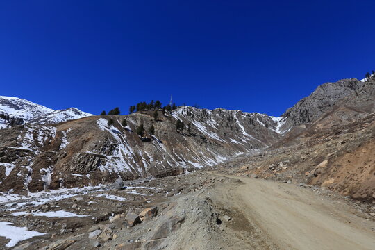 View of a rugged mountain path winds through the arid landscape, patches of snow clinging to the rocky slopes under a brilliant blue sky, Lowari Pass, Khyber Pakhtunkhwa, Pakistan.