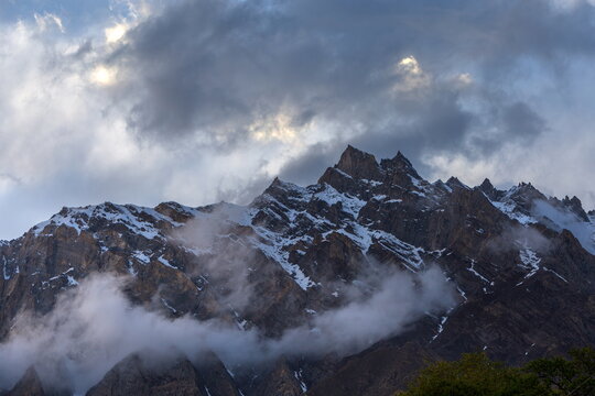 View of jagged, snow-capped peaks pierce the sky, swathed in ethereal mist and cloud veils, under a dramatic sky, Hunza, Gilgit Baltistan, Pakistan.