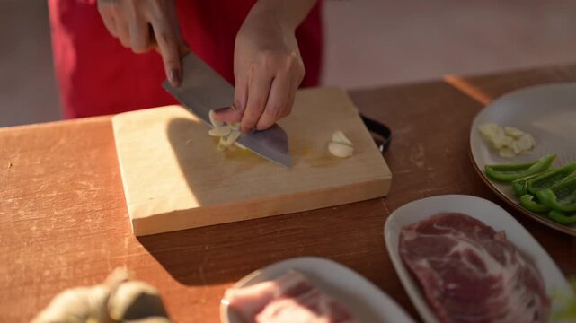 A person is cutting garlic on a wooden cutting board. There are several other items on the table, including a knife, a bowl, and a plate