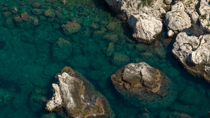 Top-down aerial detail of a rocky seabed. Crystalline emerald waters reveal large submerged boulders and jagged white rocks along the shoreline, creating a textured natural mosaic.