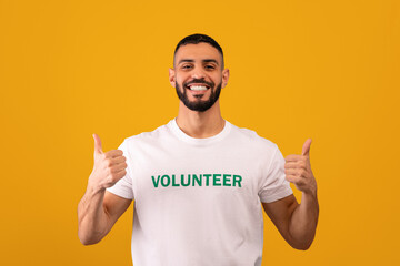 A person gives a thumbs up while wearing a shirt that reads volunteer. The background is bright yellow. The person has a beard and seems happy. This scene suggests community engagement.