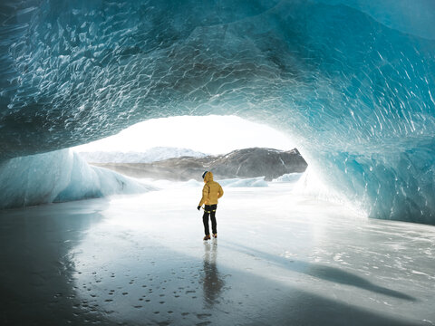 View of a lone figure in a vibrant yellow jacket stands on a frozen, reflective surface beneath a glacial ice cave, Vatnaj&Atilde;&para;kull National Park, South Iceland, Iceland.