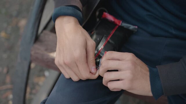 hands opening red snack wrapper on bench, closeup of mans hands tearing shiny foil packet while seated on wooden planks, navy sweater and wristband visible, autumn leaves on ground, quiet urban