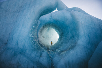 View of a lone figure standing at the end of an icy tunnel, carved by nature's hand, in a symphony of blues and whites, VatnajÃ¶kull National Park, South Iceland, Iceland.