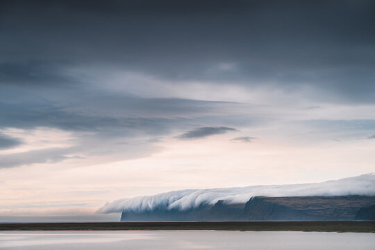 View of dramatic clouds cascade over the cliff's edge, meeting the serene waters of the Westfjords, creating a mesmerizing dance of light and shadow, Westfjords, Iceland.