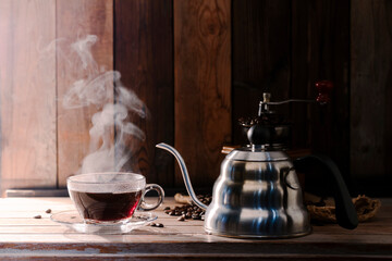 Black coffee in a cup, roasted coffee beans and a kettle on the table in a morning light atmosphere, against a backdrop of dark wood walls.