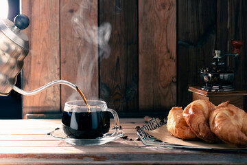 Pouring black coffee into a cup while bread is placed on the table, in the morning light, the atmosphere is warm and inviting.