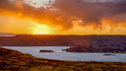 View of a sunset on Lake Titicaca with a golden sky reflecting on the water, small islands dotting the horizon, and terraced fields in the foreground, Amantani Island, Peru.