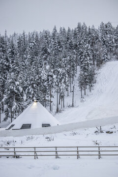 View of a snow-covered hut nestled among frosted trees, with a soft glow emanating from within, creating a serene winter wonderland scene, Rovaniemi, Lapland, Finland.