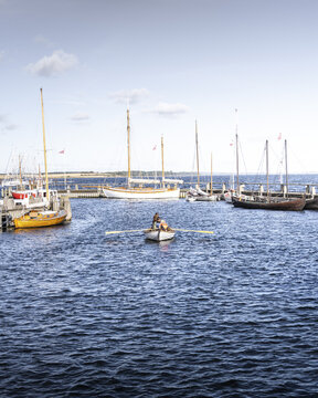 View of a lone rower cutting through the deep blue waters of the harbor, surrounded by docked sailboats under a vast sky, Roskilde, Denmark.