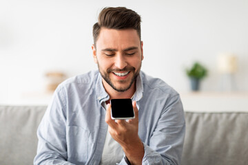 Happy handsome man recording voice message on smartphone or using virtual assistant while resting on sofa at home. Smiling guy enjoying smart technologies for communication, free space