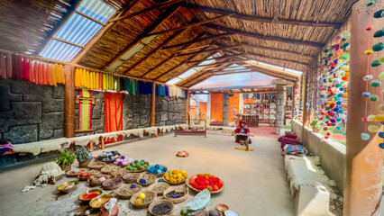 Aerial view of vibrant dyes in bowls, colorful hanging textiles, and a weaving demonstration under a thatched roof, Cusco, Peru.