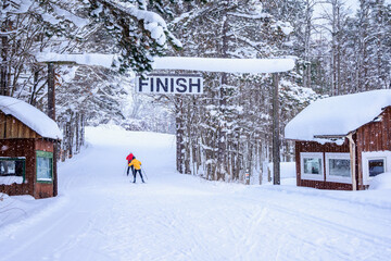 Two cross country skiers glide beneath a finish sign on a groomed trail at a nordic ski resort in winter, with snow-covered trees and small buildings on both sides.