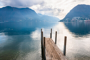 Lugano lake with Alps mountains in Lugano, Switzerland. Wooden pier on the shore of lake.