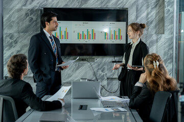 asian male business professional presenting financial chart while caucasian female team member standing and explaining analysis multiracial team collaboration in modern office meeting environment