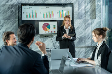 Multiracial team having strategic discussion in corporate meeting room, caucasian woman giving business presentation with charts on screen while team engages in financial planning and teamwork
