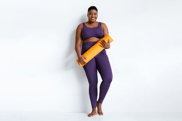 A woman smiles while standing against a white wall. She holds an orange yoga mat and is dressed in purple workout clothes. This scene captures a moment of fitness readiness.