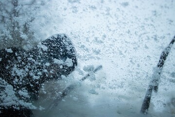 Individual in dark coat is clearing snow from a car windshield during a heavy snowstorm, showcasing the struggle against winter weather conditions and the need for vehicle maintenance