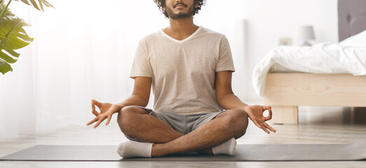 A young man sits cross-legged on a mat in a simple room. He is practicing meditation with relaxed...