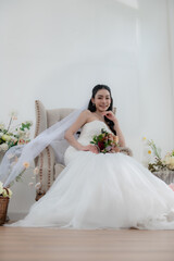 Wedding, Bride in beautiful dress sitting on sofa indoors in white studio interior, Young attractive Asian woman in wedding dress