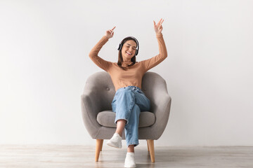 Best music app. Cool young lady sitting in armchair, listening to music in wireless headphones,...