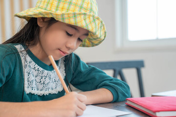 Young Asian schoolgirl writing homework at a desk with a pencil, focused and smiling, indoor learning scene showing education, creativity, and daily study routine at home or school.