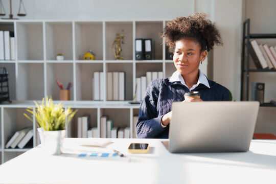 Young African American lawyer researching cases on a laptop, sipping coffee and contemplating strategy in a modern law office, focused and professional