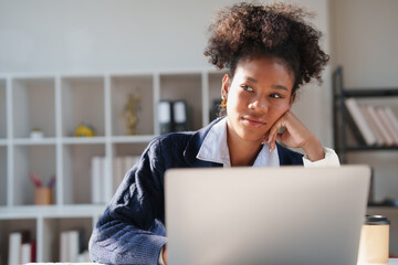 Black woman resting her head on her hand, looking away with a thoughtful expression while sitting at her desk, using a laptop, and considering new ideas