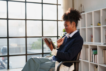 Young professional African American woman sitting comfortably in a modern office setup, concentrating while using a digital tablet with a stylus for her work or studies